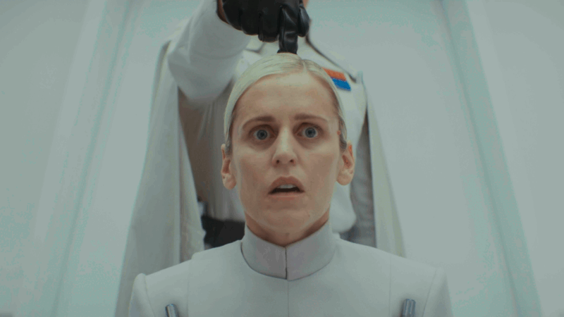 No centro da imagem está Dedra Meero sentada com um olhar assustado olhando diretamente para a câmera. Dedra é uma mulher branca com cabelo curto loiro e olhos verdes. A roupa dela é de estilo militar na cor branca, com uma gola subindo até o pescoço. Atrás dela há um homem também com roupa militar, mas utiliza uma capa e possui alguns quadradinhos coloridos localizados no lado esquerdo do peito, representando a patente maior do personagem. O personagem é Orson Krennec, com o rosto fora de foco, que está de pé e coloca um único polegar em cima da cabeça dela, com a mão dentro de uma luva preta de couro.