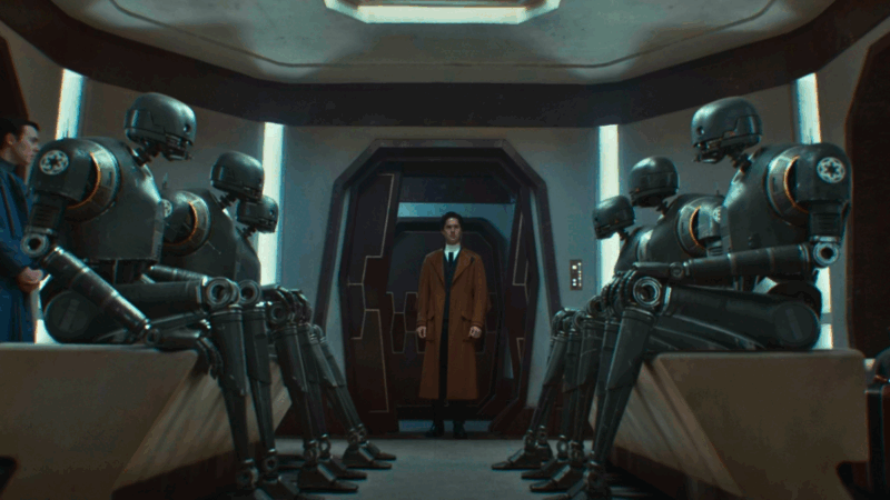 Syril Karn está no centro da imagem, em segundo plano. O personagem é um homem branco de cabelo curto e olhos azuis. Ele utiliza um casaco marrom por cima de um terno preto e gravata preta. O personagem está com o semblante assustado. Em primeiro plano, há seis droides modelo K-2 sentados, sendo 3 de cada lado. Todos olham diretamente para Syril. Os droides são pretos com uma textura metalizada. O corpo deles é humanoide, com a cabeça redonda e somente dois pontos brancos para representar os olhos.