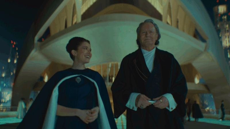 Kleya, com o mesmo vestido azul e capa azul e branca, sorri para um homem branco mais velho de peruca grisalha e expressão séria, usando um traje formal preto, parecido com um kimono, por cima de uma camisa branca. Ao fundo, aparece uma construção futurista iluminada, com arquitetura geométrica e imponente, típica de Coruscant.