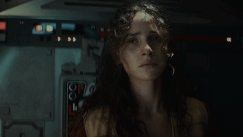 À direita da imagem está Bix, com os cabelos enrolados soltos e olha triste para alguém fora de cena. Ela está usando um colete laranja por cima de uma blusa vermelha. Ao fundo, há a parede de uma espaçonave com algumas luzes, alavancas e cabos coloridos