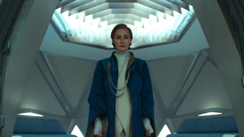 No centro da imagem há uma mulher branca mais velha com cabelo ruivo curto e penteado para a direita. É a personagem Mon Mothma. Ela utiliza um casaco azul por cima de um colete bege e uma camisa branca. Ela utiliza luvas brancas e um broche dourado com correntes que fica preso ao peito. Ao fundo, a parte de dentro um elevador com diversas luzes geométricas, formando triângulos e hexágonos brancos bastante sóbrios. Mon Mothma está com o semblante preocupado.