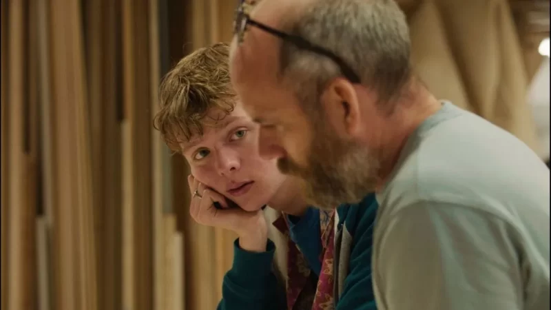 Na imagem, há um menino branco com cabelos castanhos utilizando um moletom azul. Ele está com a cabeça apoiada na mão enquanto olha um homem à sua esquerda. O homem ao seu lado é branco e calvo, com barba grisalha e vestido em um suéter cinza. 