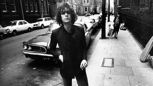 Syd Barrett, caminha por uma calçada por um bairro de Londres em uma foto em preto e branco. Ele veste terno e camisa escura, olhando diretamente para a câmera com expressão séria e um cigarro na mão esquerda. Ao fundo, há carros antigos estacionados e uma mulher carregando sacolas.