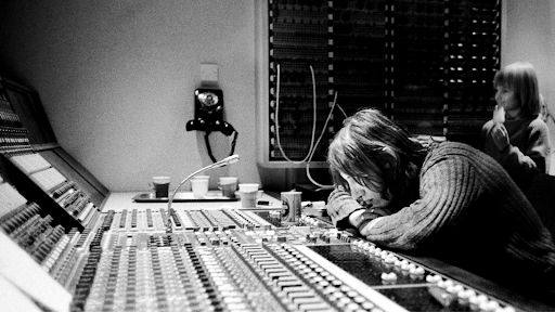 Foto preto e branca: Roger Waters debruça sobre a mesa de som do Abbey Road Studios vestindo um suéter e atrás dele um garoto, também vestindo um suéter, come um picolé enquanto observa o equipamento.