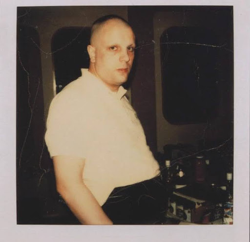 Foto polaroid: Syd Barrett, careca, obeso e sem sobrancelhas, vestindo uma camisa social branca e uma calça preta, posa de lado para a foto, encarando profundamente a câmera dentro do Abbey Road Studios.