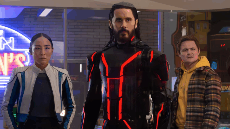 Cena do filme “Tron: Ares”. Na foto vemos os personagens Ares, Eve Kim e Seth encarando algo em sua frente. Ares, homem branco de olhos azuis e longos cabelos castanhos, veste um macacão preto com traços vermelhos. Eve Kim, mulher de descendência sul-coreana e cabelos pretos presos em um coque, usa camiseta e calças pretas e por cima veste uma jaqueta de couro branca e azul. Seth, homem pardo de cabelos castanhos claros, veste um moletom amarelo com uma jaqueta xadrez por cima. Os três estão lado à lado. Ao fundo é possível ver uma espécie de fliperama.