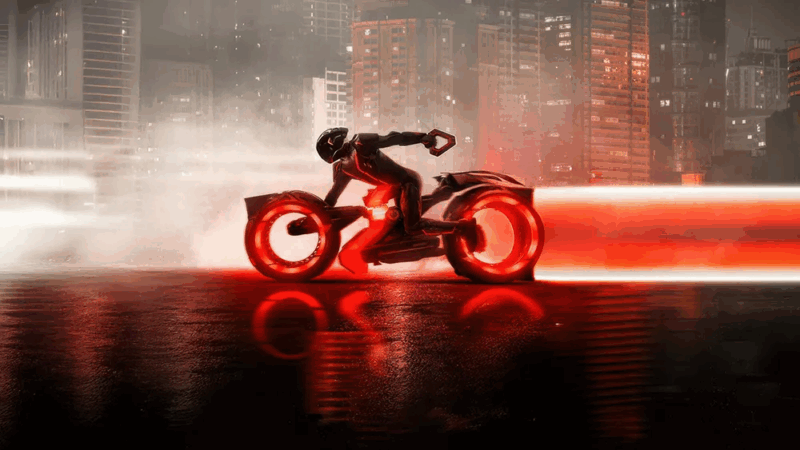 Cena do filme “Tron: Ares”. Na imagem vemos uma pessoa vestida com uma espécie de macacão e capacete ambos pretos. Ela encontra-se ao centro da foto, em cima de uma moto também na cor preta com rodas avermelhadas. Em sua mão carrega um triângulo preto e vermelho, ao fundo é possível ver grandes prédios representando uma cidade.