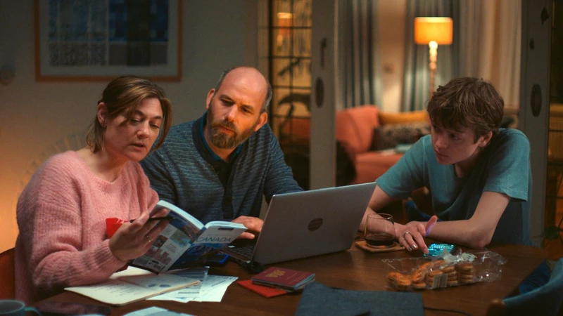 Na imagem, há uma mulher branca de cabelos castanhos presos usando um suéter rosa. Ela está segurando um livro. Ao seu lado, um homem branco calvo, de barba grisalha e ele está vestindo um suéter azul listrado. À sua frente, há um notebook aberto. No lado direito da imagem, há um menino branco com cabelos castanhos utilizando uma camiseta azul. Todos estão sentados à mesa.