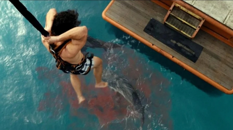 Cena do filme Animais PerigososNa imagem, do ponto de vista de cima para baixo, o personagem Moses está, de costas para a câmera, preso em um gancho, servindo de isca para tubarões. Ele está pendurado, acima do mar, balançando as pernas em desespero. Na água há sangue e um tubarão nadando. No canto superior direito da foto, há a popa de um barco. 