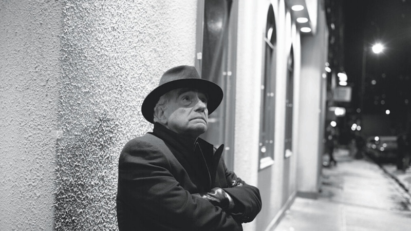 Retrato em preto e branco de Martin Scorsese, visto de perfil, já mais velho, usando chapéu e sobretudo escuro. Ao fundo, uma rua iluminada com paredes texturizadas, sugerindo um ambiente urbano.