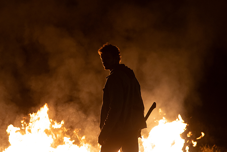 Cena do filme Love Kills.Na imagem, o personagem Marcos está de costas para a câmera, mas com o rosto virado para ela. Ele empunha um machado na mão direita. À sua frente, há muito fogo e fumaça, fazendo com a fotografia tenha bastante sombra, impossibilitando a identificação das cores de roupas do personagem, por exemplo. Marcos está vestindo calças e uma jaqueta, ele possui cabelo encaracolado. É um homem branco na faixa dos 35 anos. 