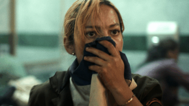 Em um close-up do filme Novembro, uma mulher loira com expressão angustiada e olhos arregalados cobre a boca e o nariz com um pano azul escuro. Ela veste uma jaqueta escura, e o fundo está desfocado com um tom esverdeado.