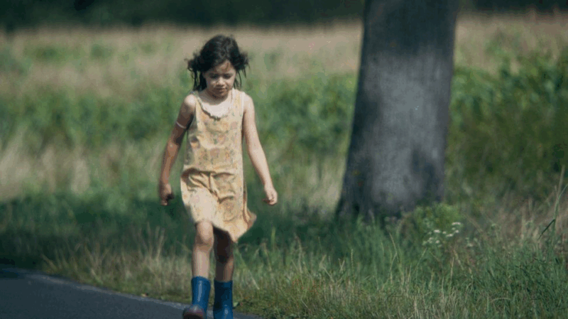Na imagem, há uma criança caminhando. Ela possui pele branca e está vestindo um vestido amarelo, botas azuis e o cabelo castanho solto. Ao fundo, é possível ver a vegetação em tons verdes e um tronco de árvore em um tom acinzentado. 