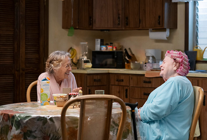 Cena do filme A Incrível Eleanor.À esquerda está Bessy, uma mulher judia, que tem os cabelos esbranquiçados e usa um óculos pequeno e veste um roupão rosa claro. Ao seu lado está Eleanor, uma senhora branca de cabelos brancos, que estão enrolados e presos com bobs. Ela veste uma roupa de pijama azul claro. Elas estão de frente uma para outra e rindo juntas. Ambas estão sentadas em uma mesa redonda na cozinha, que contém armários de madeira e utensílio na bancada ao fundo. 