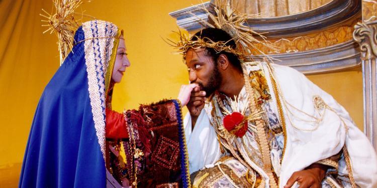 Imagem de uma cena teatral com dois personagens em trajes elaborados. Um personagem, representada pela Compadecida, veste uma túnica azul com detalhes brancos e vermelhos, segurando o rosto do outro personagem. O segundo personagem, representado por Jesus, está vestido com uma túnica branca adornada com ouro e um coração vermelho no peito, usando uma coroa de espinhos. O fundo é amarelo com ornamentos arquitetônicos, sugerindo um ambiente de palco.