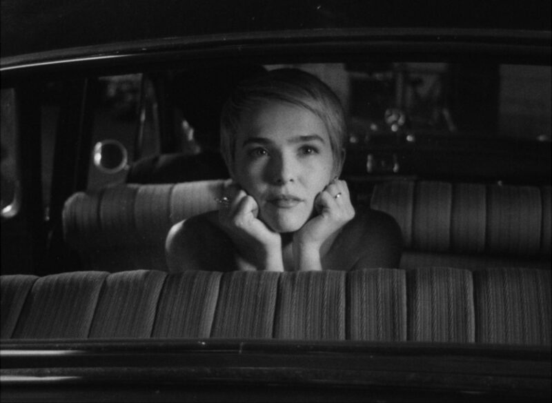 Cena do filme Nouvelle VagueNa imagem, que é em preto e branco, a personagem Jean Seberg está dentro de um carro, ajoelhada nos bancos de trás, olhando para a janela traseira, de costas para o painel do veículo. Ela está com as mãos apoiadas no rosto e olha com admiração à sua frente. Ela é uma mulher branca, na faixa dos 30 anos, de cabelos loiros e curtos, penteados para o lado. 