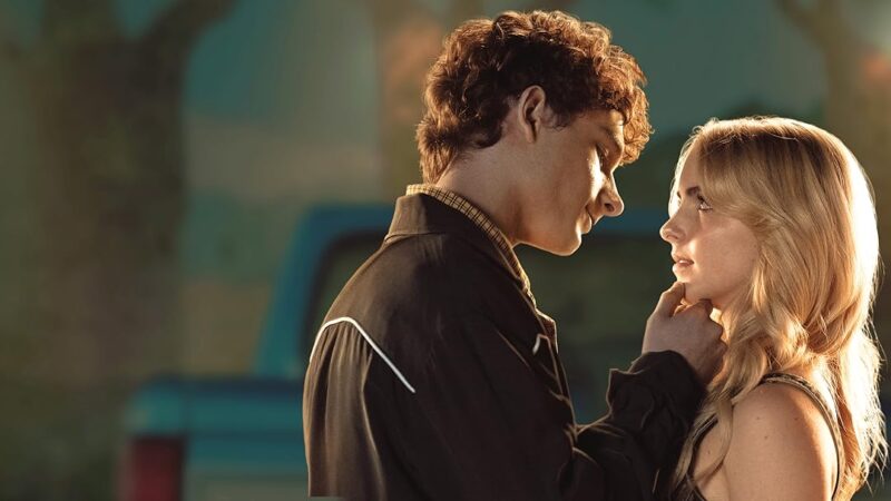 Mason Thames e Mckenna Grace estão em um momento íntimo e terno ao ar livre, diante de um veículo azul desfocado ao fundo. O rapaz, de cabelos castanhos encaracolados, veste uma jaqueta escura com detalhes claros e segura delicadamente o queixo da moça com a mão. Ele olha para ela com expressão suave e carinhosa, inclinado para a frente. A moça, loira e de cabelos longos e ondulados, encara o rapaz com olhar igualmente afetuoso, enquanto a luz dourada do entardecer ilumina seus rostos, criando uma atmosfera romântica e serena.