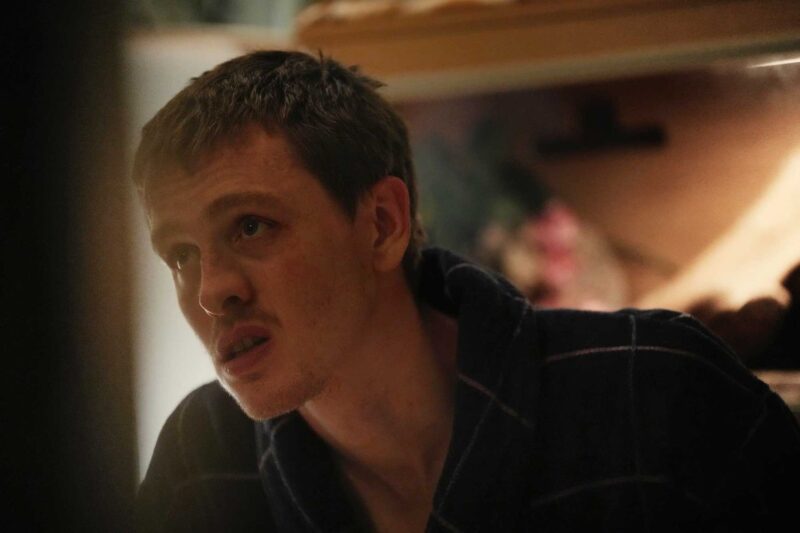 Cena do filme UrchinNa imagem, o personagem Nathan olha para alguém que está fora da cena, com uma expressão de confusão. Ele veste um roupão xadrez, na cor azul marinho. Nathan é um homem branco, de olhos claros e cabelos curtos e loiros. No rosto, usa um bigode. O cenário é de uma sala, o fundo está desfocado. 