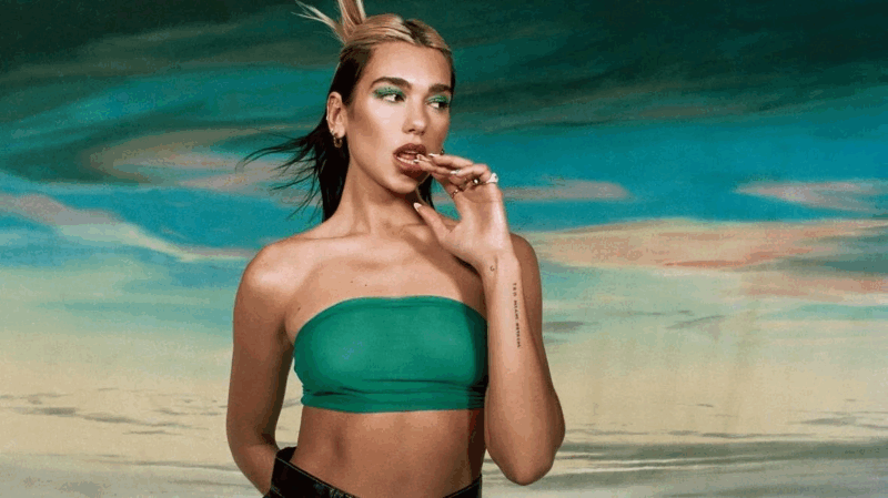 Foto da cantora Dua Lipa, uma mulher branca com cabelos louros. No registro, ela aparece com uma maquiagem verde nos olhos, semelhante ao top da mesma cor que utiliza. A cantora está com a mão esquerda na boca e olha para o lado esquerdo. Atrás dela, há uma paisagem marcada pelas cores azul, laranja e amarelo.