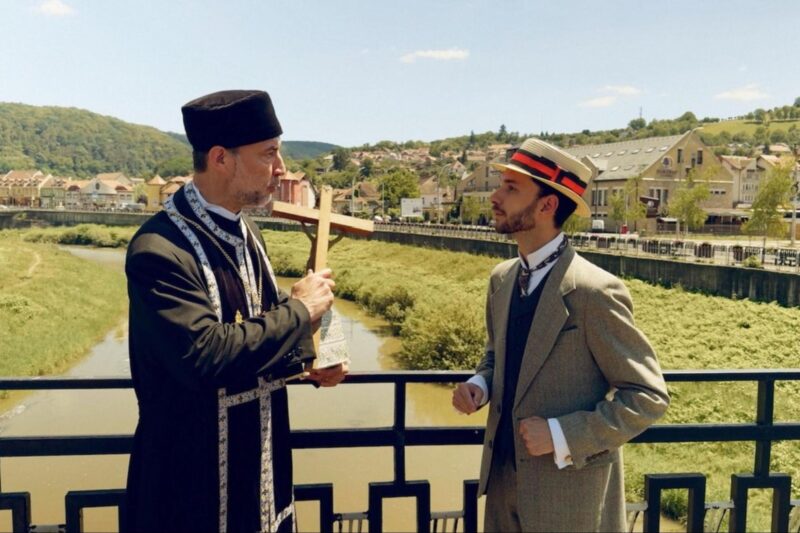 Cena do filme DraculaNa imagem, dois personagens conversam, à direita um padre e à esquerda um rapaz. Eles estão em uma ponte, de costas para a cidade. O cenário possui um rio cor marrom, gramado e casas romenas. O padre segura uma cruz com a mão direita e um pequeno baú com a esquerda, apoiado no corpo. Sua túnica é preta e possui várias faixas com cruz. Na cabeça, ele usa um chapéu religioso na cor preta. Ele possui o tom de pele claro e barba grisalha. É um homem mais velho. Já o rapaz, mais novo, veste um terno cinza, suéter azul marinho por cima de uma camisa branca e gravata por dentro. Além de um chapéu de palha com uma faixa laranja em volta. O homem também tem pele clara e barba. O dia está claro, com sol e céu azul. 