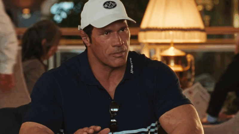 Um close-up do ator Dwayne Johnson no filme Coração de Lutador, interpretando Mark Kerr em uma cena de conversa. Ele está vestindo uma camisa polo azul-escura, que evidencia seus braços e ombros extremamente musculosos, e usa um boné de beisebol branco. Um par de óculos de sol está pendurado na gola da camisa. Com uma expressão séria e pensativa, ele olha para o lado, como se estivesse ouvindo atentamente ou falando com alguém fora da câmera. A cena se passa em um ambiente interno com iluminação suave, possivelmente um restaurante ou um clube, com um abajur e outras pessoas desfocadas ao fundo, criando uma atmosfera mais casual e íntima.