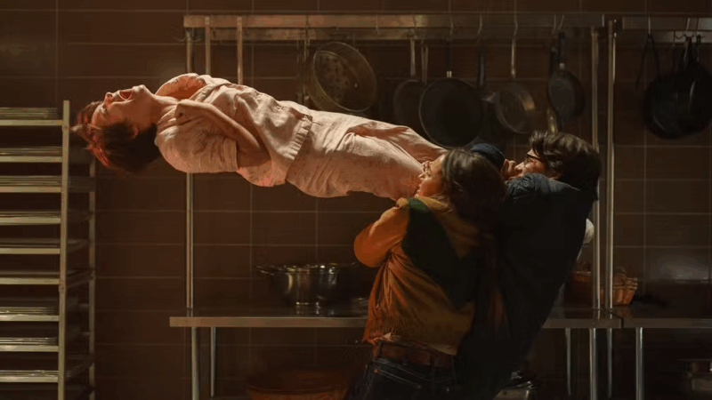 Em uma cozinha industrial, uma mulher com cabelo curto e ruivo flutua horizontalmente no ar, gritando com expressão de dor. Duas pessoas, um homem e uma mulher, tentam segurá-la pelas pernas, enquanto panelas e utensílios pendem ao fundo, reforçando a atmosfera sobrenatural da cena.