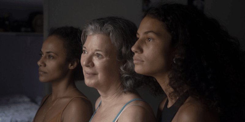Na imagem, há três mulheres de frente a uma janela. A primeira mulher possui pele negra e cabelos cacheados presos. A segunda mulher possui pele branca e cabelos grisalhos. A terceira mulher possui pele negra e cabelos cacheados longos. Todas estão contemplando o horizonte. 