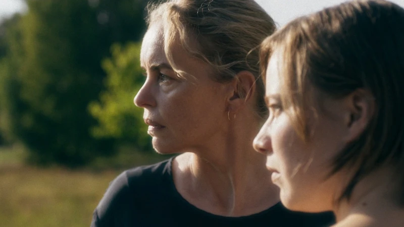 Na imagem, há duas mulheres brancas de perfil encarando o horizonte. Ambas possuem os olhos e cabelos claros. A mulher ao centro da imagem está vestindo uma camisa escura e brincos de argolas dourados. Ao fundo, é possível ver vegetações desfocadas na cor verde. 