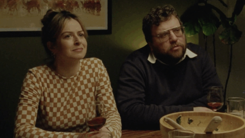 Um casal está sentado à mesa de jantar com taças de vinho pela metade. A mulher veste uma blusa xadrez em tons de marrom e branco; o homem usa suéter escuro com camisa por baixo. Eles olham para frente, atentos a algo fora do enquadramento. O ambiente tem luz quente e uma planta no canto.