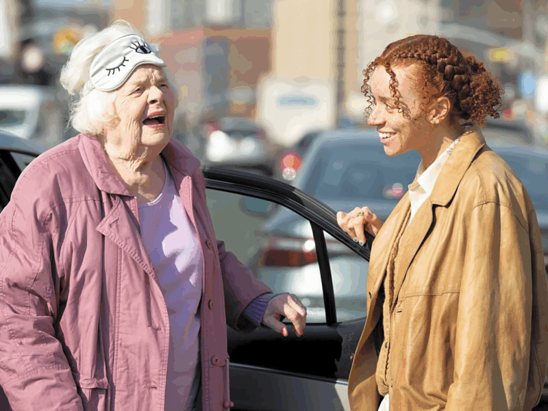 Cena do Filme A Incrível Eleanor.Eleanor, uma mulher idosa de pele e cabelos brancos e expressão alegre, vestindo um casaco rosa e uma máscara de dormir na testa. Ela está de frente a Nina, uma mulher mais jovem de cabelos cacheados ruivos e jaqueta marrom claro. Ambas estão ao lado de um carro, em um movimento de desembarque 