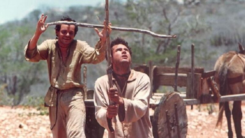 Cena em ambiente árido do sertão, com chão de terra seca e vegetação esparsa ao fundo. Dois homens vestidos com roupas simples e gastas aparecem em primeiro plano. O da esquerda, João Grilo, sorri e segura um galho de madeira em forma de cruz acima da cabeça do companheiro. O da direita, Chicó, está ajoelhado, com expressão séria e olhar voltado para cima, também segurando um pedaço de madeira em formato de cruz. Atrás deles há uma carroça de madeira puxada por um animal, sugerindo um contexto rural e de pobreza.