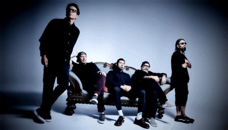 Cinco pessoas brancas posam diante de um sofá antigo com entalhes de madeira e estofado estampado. Duas estão sentadas e três em pé ao redor, todas vestindo roupas escuras e modernas. O fundo em tom azul cria contraste com o mobiliário clássico e a postura descontraída do grupo.