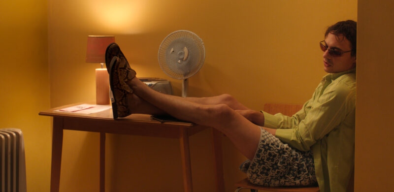 Cena do filme UrchinNa imagem, o personagem Mike está sentado em uma cadeira, com as costas apoiadas na parede e pernas em uma mesa. Ele está à direita da fotografia, vestindo camisa de botões verde clara, óculos escuros, shorts com estampas de cruz na tom verde e mocassim de pele de cobra. Ele está com as costas apoiadas no canto da sala, em uma parede, enquanto as costas da cadeira estão na parede da frente. Na mesa de madeira, há um ventilador branco de hélices azuis, um rádio grande, um abajur aceso e uma folha. No canto inferior esquerdo há um aquecedor. As paredes são amarelas. Mike é um rapaz branco, na faixa dos 30 anos, de cabelos castanhos claros e barba por fazer. 