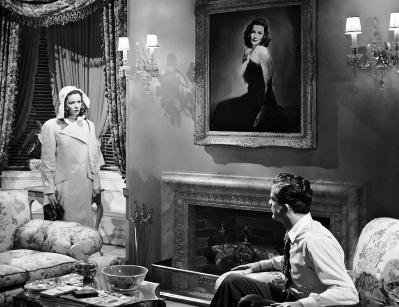 Cena de Laura. O filme é em preto e branco. O cenário é o interior de uma casa. À direita está o detetive McPherson, usando uma camisa branca com gravata. Ele está sentado em uma poltrona florida de lado para a câmera. Ele olha para a verdadeira Laura que está à esquerda da imagem. Ela usa um sobretudo claro e um chapéu da mesma cor. Seu rosto demonstra irritação e olha diretamente para McPherson. A iluminação a deixa em evidência. Ao centro, entre os dois, está a pintura, que ganha aspectos ainda mais dramáticos por causa da cena.