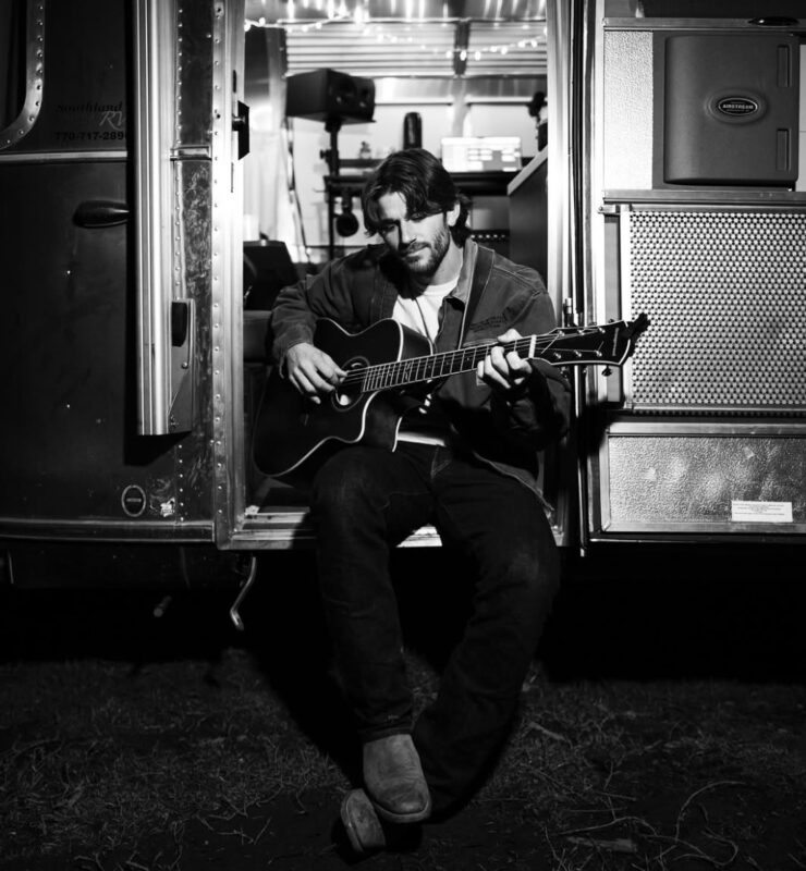 Fotografia postada no Instagram de Jonah Kagen. A imagem está em preto e branco. Um homem está sentado na porta aberta de um trailer metálico. Ele está com a cabeça baixa tocando um violão preto. Ele usa uma camiseta branca, jaqueta jeans, calça escura e botas marrons. O interior do trailer está iluminado suavemente e cria um contraste com o exterior escuro.