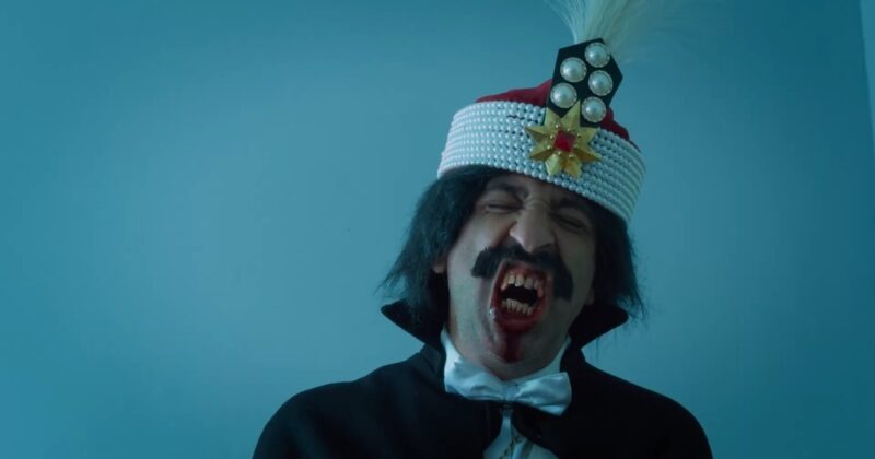 Cena do filme DraculaNa imagem, um dos personagens que representa o Dracula ao longo do filme, aparece com uma expressão de prazer, enquanto recebe um sexo oral. Ele está com os olhos fechados e a boca ensanguentada e aberta, mostrando os dentes afiados. Seus cabelos são na altura do pescoço. Também usa um bigode. Dracula está vestindo uma camisa de botões branca, gravata borboleta da mesma cor e uma capa preta com gola alta. Na cabeça, ele usa um chapéu vermelho com adorno. Na ponta, é vermelho, enquanto a borda é enfeitada por pérolas. No centro, há uma estrela dourada de 8 pontas com uma pedra vermelha e quadrada no centro, em volta dela há outras pedras redondas douradas, mas menores. Acima da estrela, há um pentágono preto com cinco pedras redondas na cor branca, assim como uma pena branca anexada. 