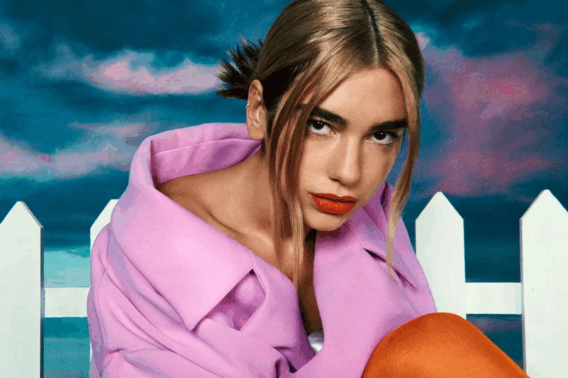 Foto da cantora Dua Lipa, uma mulher branca de cabelos louros. No registro, ela veste um casaco rosa e olha para frente. Ela está com o cabelo preso e com duas mechas soltas na frente do rosto.