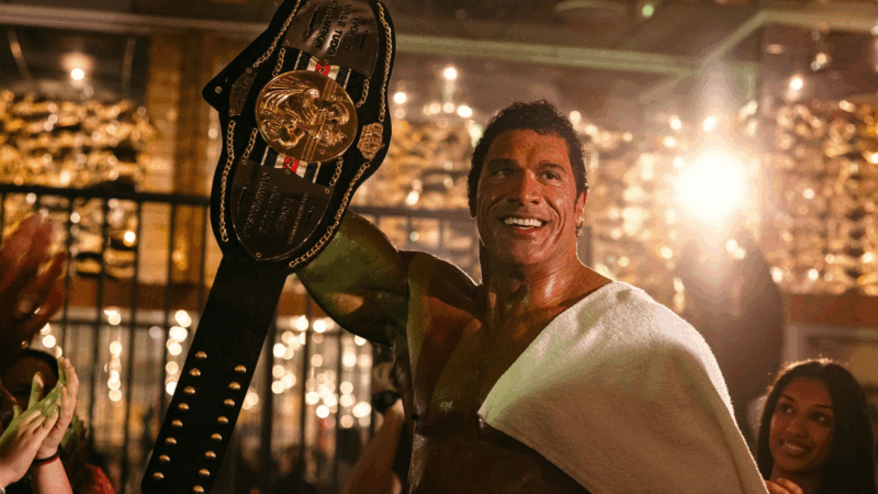 Em uma cena do filme Coração de Lutador, o ator Dwayne Johnson, interpretando o lutador Mark Kerr, celebra uma vitória. Ele está em pé, sem camisa, com o corpo musculoso e brilhando de suor. Uma toalha branca está casualmente jogada sobre seu ombro esquerdo. Com a mão direita, ele ergue um grande cinturão de campeão, que tem uma faixa preta e uma placa central dourada com detalhes em relevo. Dwayne Johnson olha para cima e para o lado com um sorriso largo e radiante, expressando pura alegria e triunfo. O fundo é caloroso e festivo, preenchido com luzes douradas e desfocadas que criam um efeito brilhante (bokeh). À esquerda, mãos de pessoas aplaudem, e à direita, o rosto de uma mulher sorrindo é parcialmente visível, completando a atmosfera de comemoração.
