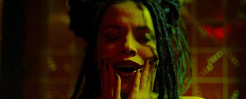 Cena do filme Love Kills.Na imagem, com cores em tons de verde neon, a personagem Helena está com as duas mãos nas bochechas com expressão de prazer. Ela é uma vampira e se delicia com sangue na boca, que está aberta, mostrando as presas. Ela está de olhos fechados e a fotografia fecha o plano em seu rosto. Ao fundo, em desfoque, há uma parede laranja. Helena é uma mulher negra, na faixa dos 35 anos, de cabelos dread na cor preta. Suas unhas estão pintadas em tom escuro. 