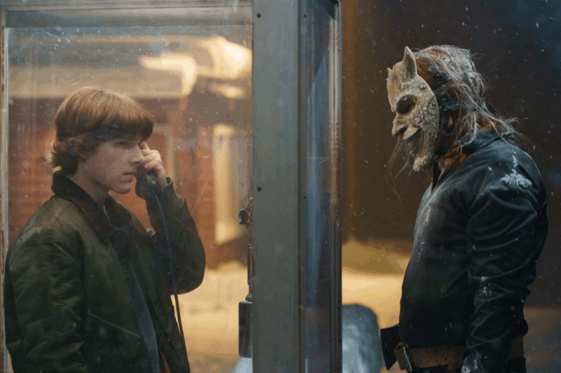 Um adolescente de cabelo castanho e jaqueta verde está dentro de uma cabine telefônica, segurando o telefone próximo ao ouvido e olhando atentamente para um homem do lado de fora. O homem usa uma máscara pálida e rachada com chifres, cabelos longos e roupas escuras, observando o garoto sob uma luz fria e tensa.