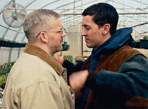 Texto alternativo: Cena do filme À Paisana. Na imagem, há um homem branco de cabelos grisalhos, que usa uma jaqueta bege e um homem branco de cabelos castanhos escuros, que veste um moletom azul. Eles estão se beijando.