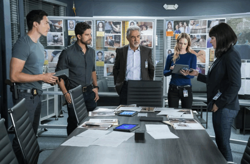 Em um escritório, há 5 pessoas. Da esquerda para a direita: um homem alto com cabelo preto, um homem mais baixo com cabelo preto e barba, um homem de cabelos brancos com roupas sociais, uma mulher loira com blusa azul e uma mulher de cabelo preto e franja. A mulher loira olha para um tablet que está em sua mão. O primeiro homem também segura um tablet, mas olha para a mulher de franja, que segura um celular. Todos estão com expressões sérias. No fundo da sala há dois painéis com fotos dos crimes. Na mesa, há arquivos dos casos, fotos e tablets.