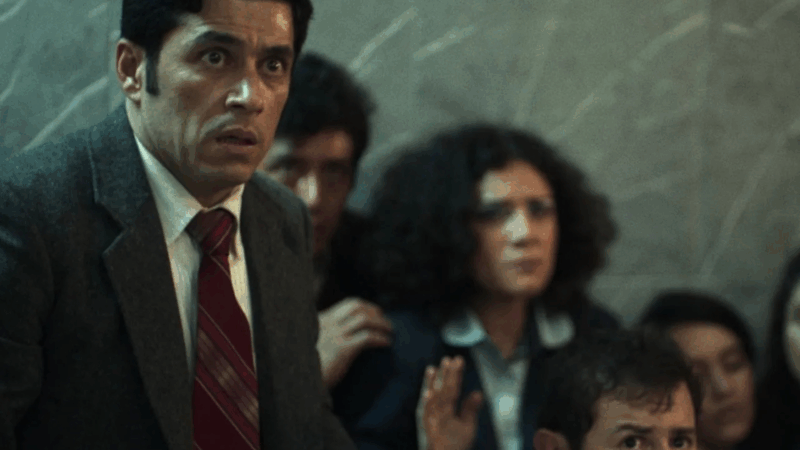 Em uma cena do filme Novembro, um homem de terno cinza e gravata vermelha listrada olha para o lado com uma expressão de choque e alarme, com os olhos arregalados. Ele está em um local cheio, com outras pessoas ao fundo, incluindo uma mulher de cabelos cacheados que observa com medo.