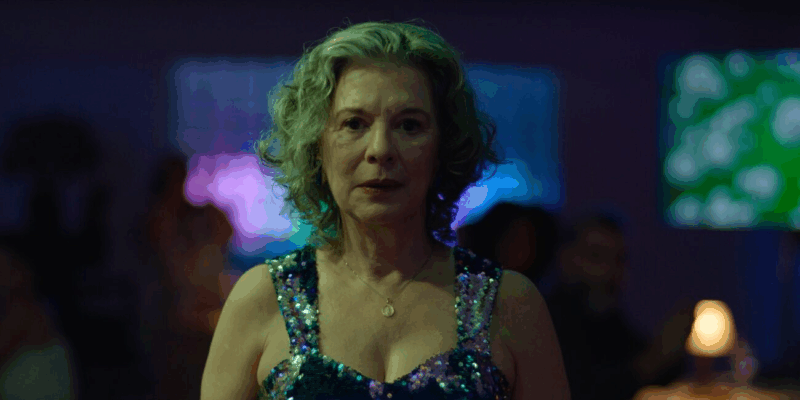 Na imagem, há uma mulher branca de cabelos grisalhos. Ela está com o rosto maquiado e usando um vestido colorido. Em seu pescoço, há um colar dourado. 