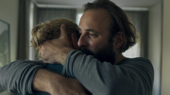 Na imagem, há um homem e uma mulher abraçados. O rosto dela está escondido no ombro dele. A mulher possui cabelos loiros e o homem possui cabelo e barba castanhos. Ele está vestindo um moletom cinza e, em seu rosto, há uma expressão de tristeza. Ao fundo, é possível ver a luz de uma janela. 