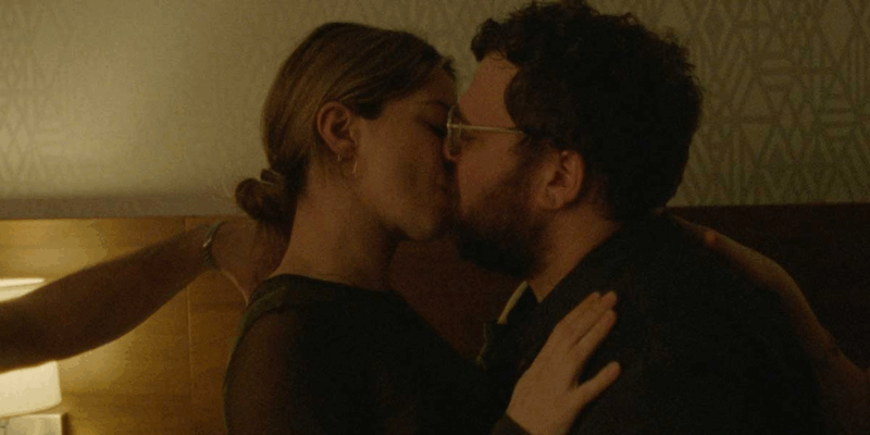  Um casal se beija em um quarto com iluminação amarelada. A mulher, de cabelo preso, segura o rosto do homem, que usa óculos. Ao fundo, há uma parede com padrão geométrico e parte de um abajur. A cena é íntima e filmada de perto.