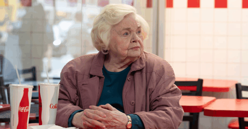 Cena do filme A Incrível Eleanor.Na imagem, aparece Eleanor, uma mulher branca, idosa, de cabelos brancos, ​​que está sentada em uma lanchonete, usando um casaco rosa e uma blusa verde-azulada. Ela está com as mãos apoiadas sobre a mesa e olha para o lado parecendo pensativa. À sua frente há dois copos grandes de refrigerante. Ao fundo, há mesas e cadeiras vermelhas e pretas. 