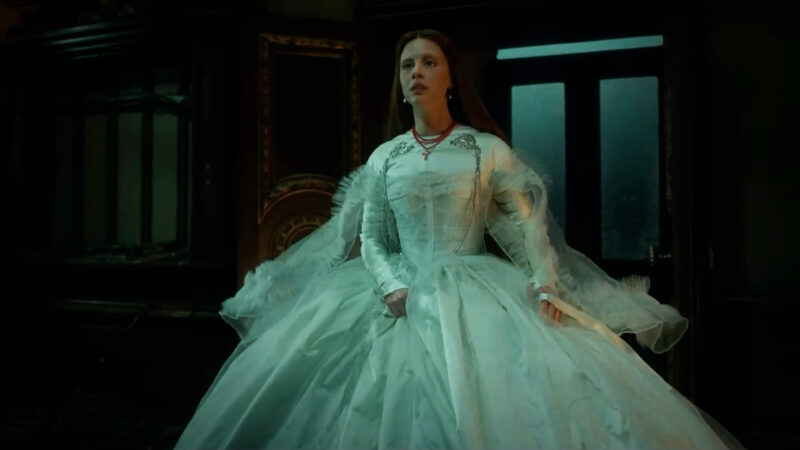 Cena do filme Frankenstein Na imagem, Elizabeth olha com surpresa para á frente. Ela veste um vestido branco de noiva e um terço vermelho no pescoço. O vestido é bem volumoso e possui detalhes em prata na região da clavícula. A cena é noturna, se passa num quarto, atrás dela há uma porta com janelas de vidro. Elizabeth é uma mulher branca, na faixa dos 35 anos, de cabelos longos na cor ruiva. 