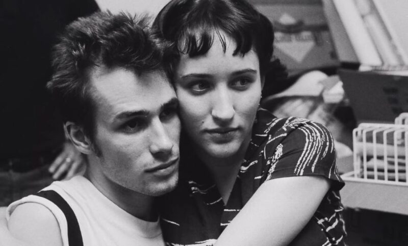 Cena do filme It’s Never Over, Jeff BuckleyNa imagem, em preto e branco, Jeff Buckley e Rebecca Moore estão abraçados e sérios. Ela, à direita, está sentada no colo do cantor, usando uma camiseta de listras com o colarinho aberto. Possui pele clara, cabelos escuros amarrados e com a franja dividida ao meio. Já Jeff é um homem branco, de cabelos escuros e lisos, penteado de forma arrepiada. Está vestindo uma regata branca com uma faixa preta no ombro. 