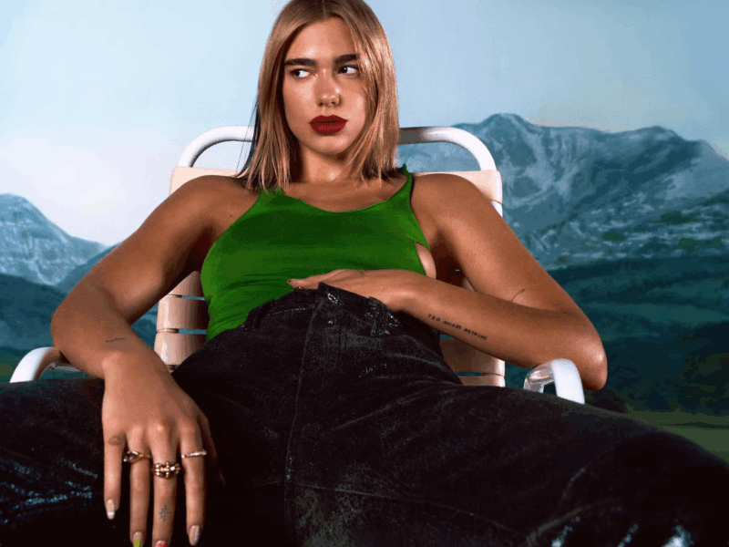 Foto da cantora Dua Lipa, uma mulher branca com cabelos louros. No registro, ela aparece sentada em uma cadeira de praia. Ela veste uma blusa regata verde e uma calça jeans preta. Ela possui anéis na mão direita e está com a mão esquerda dentro da calça. Ela olha para o lado direito e usa um batom da cor vermelho escuro. Atrás da cantora, há espécies de montanhas com tons de azul.
